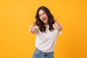 Woman with brown hair winking and giving two thumbs up smiling in front of yellow background